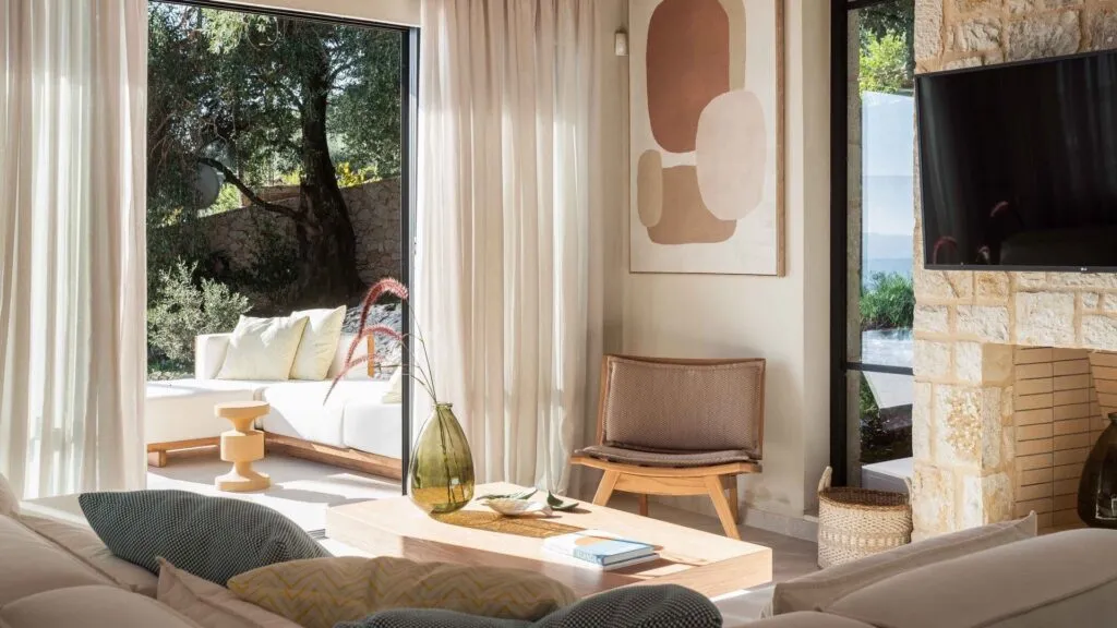 Inside luxury villa San stefano. Muted beige and brown interior with large couch and chair. muslin soft curtains over window looking out into courtyard. Photograph taken in Greece, Corfu